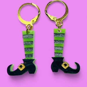 Pode incluir: Um par de brincos de bota de bruxa com pernas listradas verdes e prateadas, botas pretas e fivelas douradas. Os brincos são feitos de argila polimérica e têm ganchos dourados.