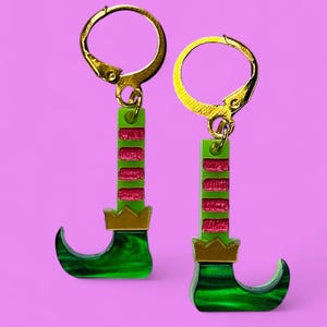 May include: A pair of festive elf leg earrings. The earrings feature green and red striped legs with gold-colored accents, including the hook and the crown on the shoe. The earrings are set against a pink background.