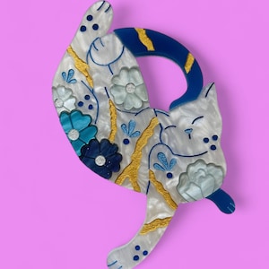 May include: A white cat brooch with blue and gold floral details. The cat is curled up with its tail wrapped around its body. The brooch is made of resin and has a shiny finish.