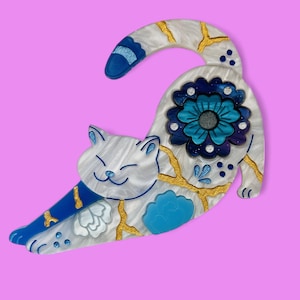 May include: A white ceramic cat brooch with blue and gold accents. The cat is stretching and has a flower on its back.