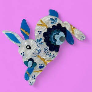 May include: A decorative bunny brooch in white, blue, and gold. The brooch features a stylized rabbit design with blue ears, feet, and floral accents. The bunny's body has a marbled white pattern with gold accents. The brooch is set against a pink background.