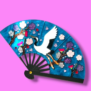 Op de afbeelding: Decoratieve waaier met een turquoise achtergrond, met een witte kraanvogel en bloemaccenten. De waaier heeft een zwarte handgreep en is versierd met roze, witte en paarse bloemen. Het ontwerp van de waaier doet denken aan Japanse kunst.