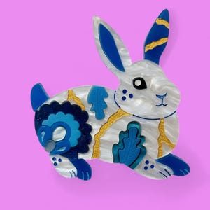 May include: A white rabbit brooch with blue and gold details. The rabbit has a blue ear, a gold ear with blue stripes, and a blue and gold patterned body.