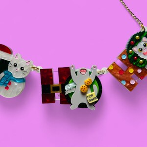 Pode incluir: Um colar de corrente dourada com um gato branco usando um chapéu de Papai Noel, um gato branco usando um cachecol azul, um gato branco segurando biscoitos e um gato branco usando uma coroa. O colar soletra "HO HO" em letras coloridas.