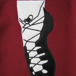 May include: A black and white embroidered patch of a climbing shoe with white laces and a black sole.