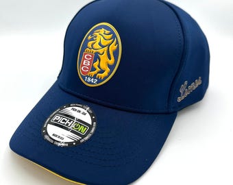 May include: A navy blue baseball cap with a curved brim and a yellow under brim. The cap features a gold and yellow lion crest with the text "CBC 1842". The word "Lions" is embroidered on the side. A "PICH-ON" sticker is on the brim.