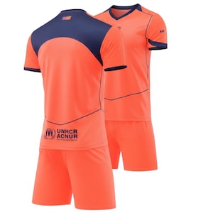 May include: Two-piece soccer uniform set in bright orange with navy blue accents. The jersey features a V-neck and a navy blue shoulder panel. The shorts are solid orange. The UNHCR ACNUR logo is printed on the jersey.