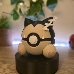 Op de afbeelding: Een zwart, wit en bruin Pokemon-figuur van een slapende Snorlax. Het figuur staat op een zwarte basis.