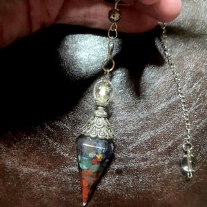 Op de afbeelding: Een zilverkleurige pendel met een facetgeslepen kristal aan het einde van een ketting. De pendel heeft een decoratieve zilveren bovenkant en een kleurrijk, gelaagd edelsteenlichaam. De edelsteenkleuren zijn rood, oranje, geel, groen, blauw en paars.