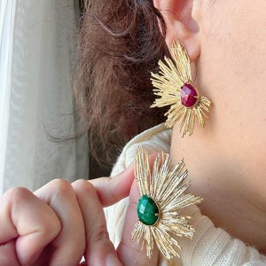 Könnte beinhalten: Ein Paar goldene Ohrringe mit einem Sternburst-Design und einem grünen und roten Edelstein in der Mitte.