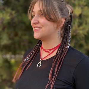 May include: A woman wearing a black t-shirt and a red necklace with a crescent moon pendant. Her hair is styled with braids and earrings. The background is a blurred green.
