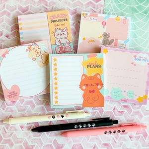 Può includere: Una collezione di post-it e penne colorate. I post-it presentano illustrazioni di gatti e frasi come "Crazy Projects" e "Boo-tiful". Sono visibili tre penne, con disegni di impronte di zampe. Lo sfondo ha un motivo rosa e bianco.