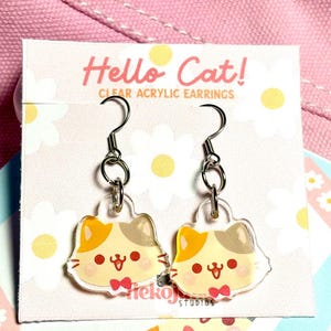 Puede incluir: Pendientes de acrílico transparente con un diseño de gato con detalles en naranja, beige y gris, y una pajarita roja. Los pendientes están sobre una tarjeta blanca con el texto "Hello Cat! CLEAR ACRYLIC EARRINGS". El fondo es rosa.