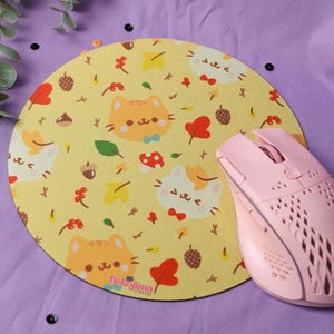 Op de afbeelding: Een gele muismat met een herhalend patroon van schattige cartoonkatten en herfstbladeren. De muismat is rond en heeft een roze computermuis erop.