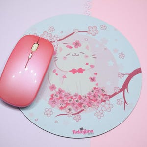 Pode incluir: Um tapete de rato rosa com um design de gato branco. O gato está rodeado de flores de cerejeira cor-de-rosa e o texto "Neko-jima Studios" está no canto inferior direito.