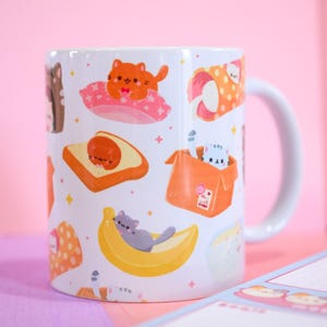 Può includere: Tazza in ceramica bianca con un motivo ripetuto di gatti dei cartoni animati in varie pose, come su un cuscino, in una scatola e su una banana. La tazza ha un manico bianco ed è ambientata su uno sfondo rosa.