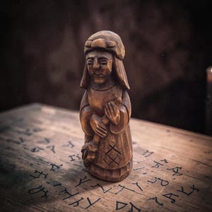 Puede incluir: Una figura de madera tallada a mano que representa a una persona con gorro y túnica larga, sosteniendo un bastón. La pieza muestra marcas de cincel visibles y un símbolo geométrico grabado en la parte frontal de la prenda de madera marrón.