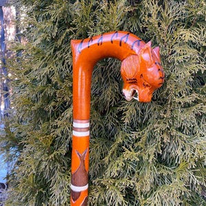 Puede incluir: Un bastón de madera con una cabeza de tigre tallada en la parte superior. El bastón está pintado de naranja y marrón con rayas blancas.
