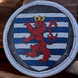 May include: A white round shield with a black border and a blue and white striped background. A red lion with a gold crown is in the center of the shield.