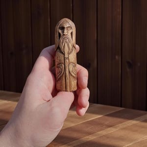 Könnte beinhalten: Eine geschnitzte Holzfigur eines bärtigen Mannes mit Kapuzenmantel. Die Figur hält einen Stab, und zwei Vögel sind auf dem unteren Teil geschnitzt. Das Holz hat eine natürliche, hellbraune Farbe.