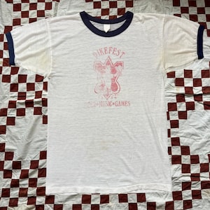 Puede incluir: Camiseta blanca con cuello y ribetes de manga azul marino. La parte delantera presenta un gráfico rojo descolorido con el texto "BIKEFEST" encima de un diseño y "1989" y "MUSIC GAMES" debajo.