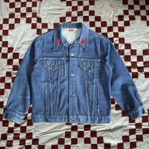 May include: A blue denim jacket with red embroidered hearts on the collar. The jacket has a button closure and two front pockets.