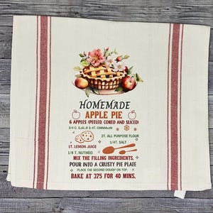 Könnte beinhalten: Cremefarbenes Küchentuch mit roten vertikalen Streifen und einer Grafik von Apfelkuchen. Die Grafik zeigt einen Kuchen, Äpfel und Blumen, mit einem Rezept für hausgemachten Apfelkuchen. Der Text enthält Zutaten und Backanweisungen.