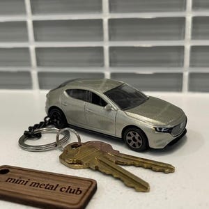May include: A silver model car keyring with a set of keys and a wooden tag that reads "mini metal club". The car is a hatchback style with black wheels.