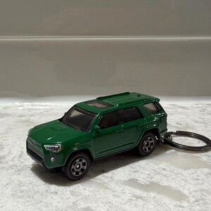 May include: A dark green toy SUV keyring featuring a detailed exterior with a sunroof, black wheels, and a metal ring attached to the rear. The miniature vehicle has a glossy finish and realistic headlights, capturing the design of a modern sport utility vehicle.