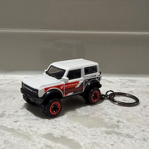 Puede incluir: Un llavero de vehículo de juguete Ford Bronco blanco con guardabarros negros, gráficos laterales rojos y negros, y ruedas con detalles rojos. La palabra BRONCO está impresa en rojo en el lateral y en negro en la parrilla frontal, unido a una argolla.
