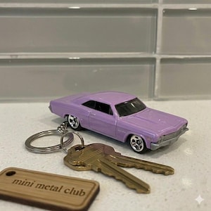 Könnte beinhalten: Ein Schlüsselanhänger mit einem lavendelfarbenen Spielzeugauto, einem silbernen Ring und zwei goldenen Schlüsseln. Ein kleines Holzschild mit dem Text "mini metal club" ist ebenfalls angebracht. Das Auto ist ein klassisches Modell.