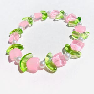May include: A pink and green beaded bracelet with flower-shaped charms. The bracelet is made with alternating pink and green beads, with white pearl beads interspersed throughout.