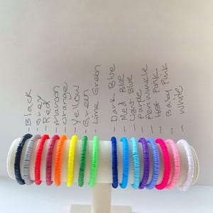 May include: A display of colorful beaded bracelets in various colors, including black, grey, red, maroon, orange, yellow, green, lime green, dark blue, medium blue, light blue, purple, periwinkle, hot pink, baby pink, and white. The bracelets are arranged on a white stand.