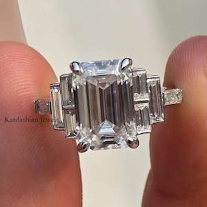 May include: A silver ring featuring a large, rectangular emerald-cut central gemstone, with smaller baguette-cut stones on either side. The ring is held between fingers. The text "Kardashian Jewels" is visible on the ring.