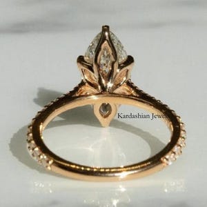 May include: Rose gold engagement ring with a pear-shaped diamond center stone. The band is encrusted with small diamonds. The ring is set against a white marble surface. The text "Kardashian Jewel" is visible on the ring.
