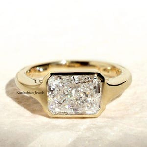 May include: A gold ring featuring a large, rectangular, clear gemstone. The ring has a smooth, polished band and a bezel setting. The ring is set against a neutral background. The brand name "Kardashian Jewels" is visible.