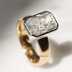 May include: A gold ring featuring a large, rectangular-cut diamond set in a platinum bezel. The band is gold and polished, with the text "Kardashian Jewels" engraved on the side. The diamond sparkles brightly, reflecting light. A luxury jewellery item.