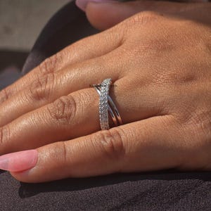 May include: A silver ring with a crisscross design, set with small, sparkling stones. The ring is displayed on a hand with a light skin tone and long, pink nail polish.