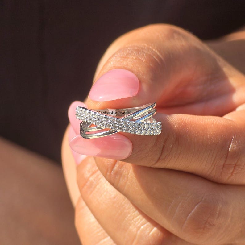 May include: A silver-coloured criss-cross ring with a row of small, clear stones set across the top band. The ring is held in a hand with pink nail polish. The brand name "Kardashian Jewels" is visible on the ring.