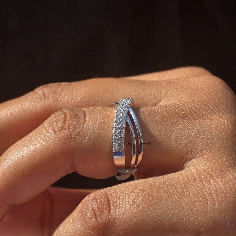 May include: A silver ring with a double-band design. One band is encrusted with small, clear gemstones, while the other two bands are smooth and polished. The ring is shown on a finger against a blurred background.