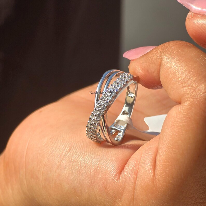 May include: A silver ring with a crossover design. One band is set with small, clear stones. The ring is held in a hand with pink nail polish. The text "Kardashian Jewels" is visible.