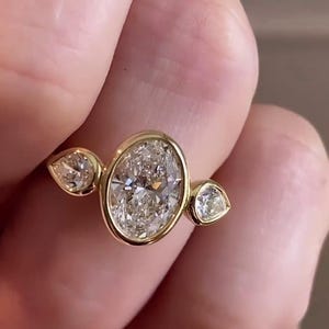 May include: A gold ring with a large oval-shaped diamond and two smaller pear-shaped diamonds on either side.