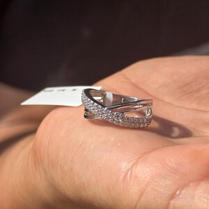 May include: A silver ring with a crisscross design, adorned with small, sparkling stones. The ring's design is highlighted against a neutral background, showcasing its intricate details and elegant style. The ring is a piece of jewellery.