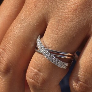 May include: A silver ring with a crisscross design, featuring a row of small, sparkling stones. The ring is worn on a finger, showcasing its elegant and modern style. The ring is a piece of jewellery.