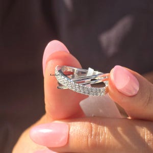 May include: A silver ring with a crisscross design, set with small, sparkling stones. The ring is held up close, highlighting the intricate details and the light reflecting off the stones. The ring is a piece of jewellery.