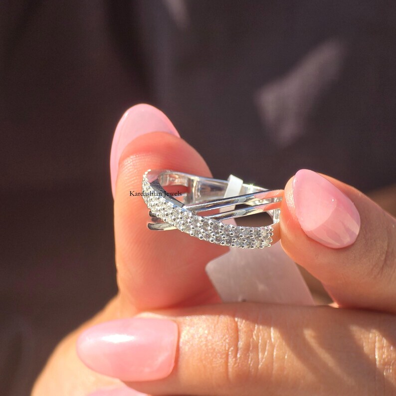 May include: A silver ring with a crisscross design, set with small, sparkling stones. The ring is held up close, highlighting the intricate details and the light reflecting off the stones. The ring is a piece of jewellery.