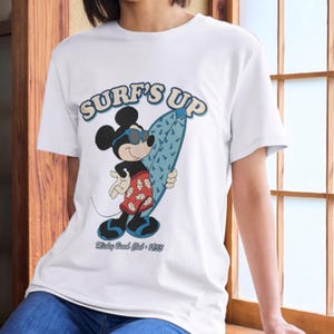 May include: White t-shirt featuring Mickey Mouse in sunglasses holding a blue surfboard. The shirt has the text "SURF'S UP" in blue and the text "Mickey Beach Club - 1955" in black.