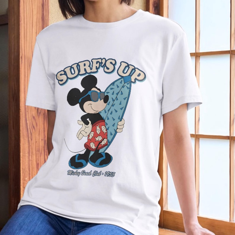 May include: White t-shirt featuring Mickey Mouse in sunglasses holding a blue surfboard. The shirt has the text "SURF'S UP" in blue and the text "Mickey Beach Club - 1955" in black.