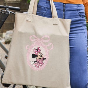 May include: Beige canvas tote bag featuring a pink oval frame with Minnie Mouse in a pink polka dot dress and bow. The frame is adorned with a pink bow and ribbons. The bag has two handles and is held by a person.