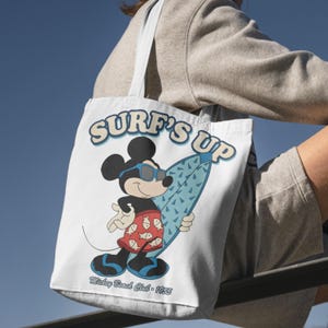 May include: White tote bag featuring Mickey Mouse in sunglasses, holding a blue surfboard. The words "SURF'S UP" are printed above the character, with "Mickey Beach Club - 1955" below. The bag has light blue straps.